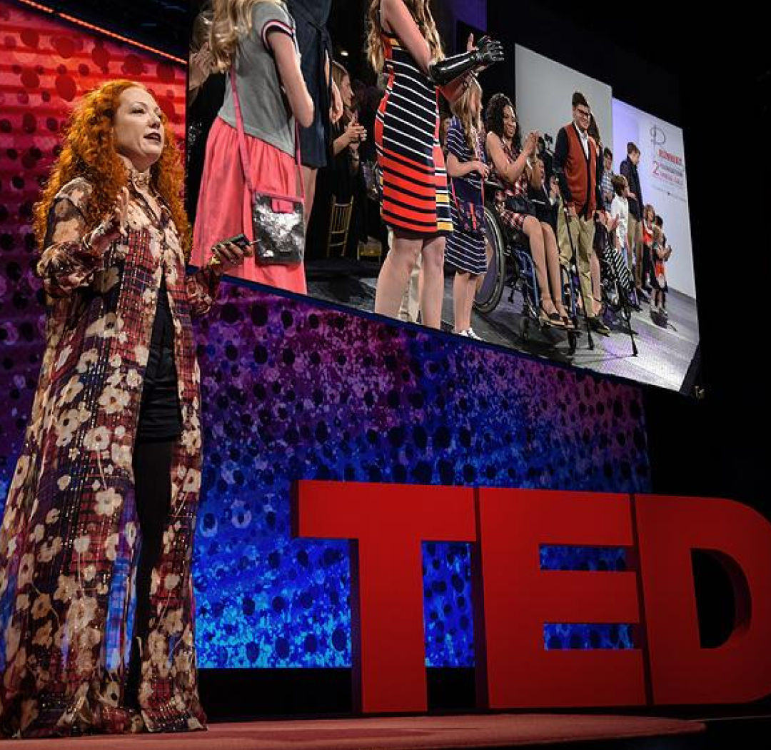 Mindy Scheier, a woman with long red curly hair wearing a long floral top over black clothes on a stage with large red letters spelling TED and a screen in the background.