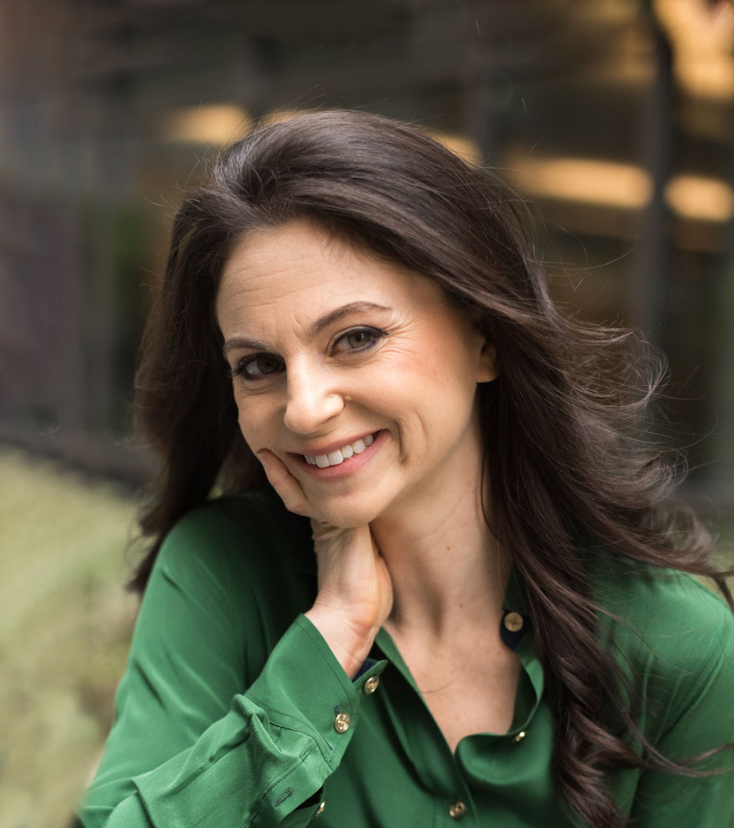 Woman with long brown hair smiling with hand under her face wearing a green button-up shirt.