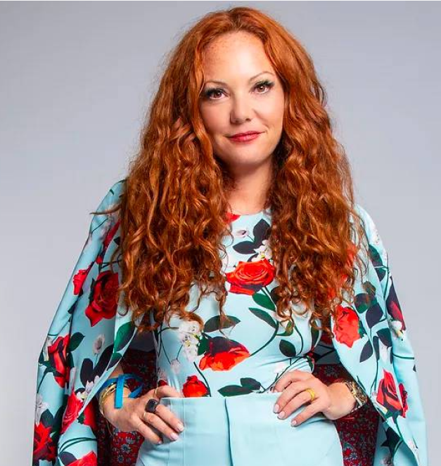 Mindy is a woman with long curly red hair wearing light blue pants with a light blue top and cape that has red flowers on it. She is posing with her hands on her hips and a slight smile.