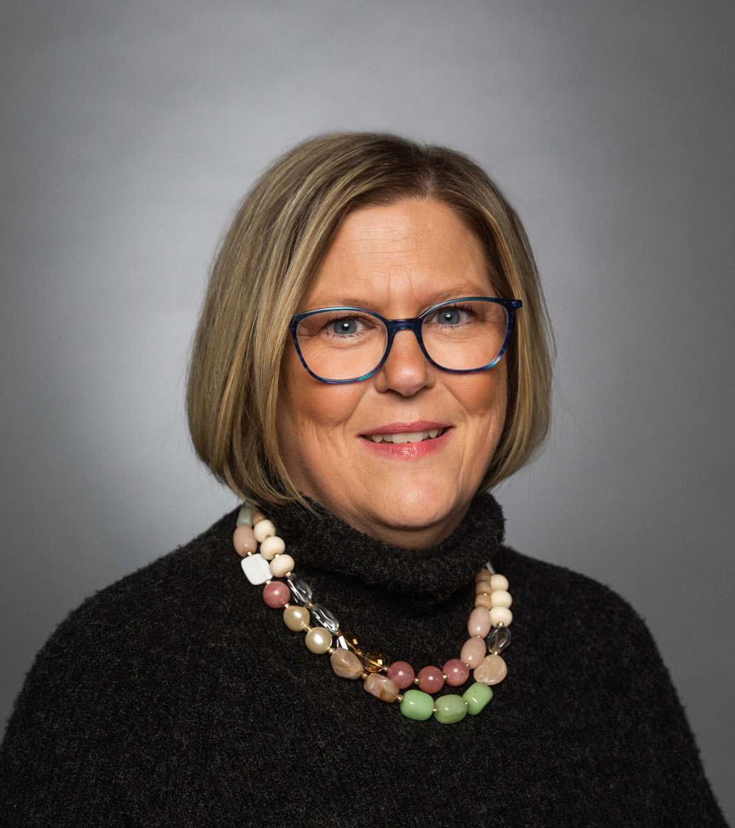 Woman with short blond-brown hair with glasses wearing a beaded necklace and black shirt