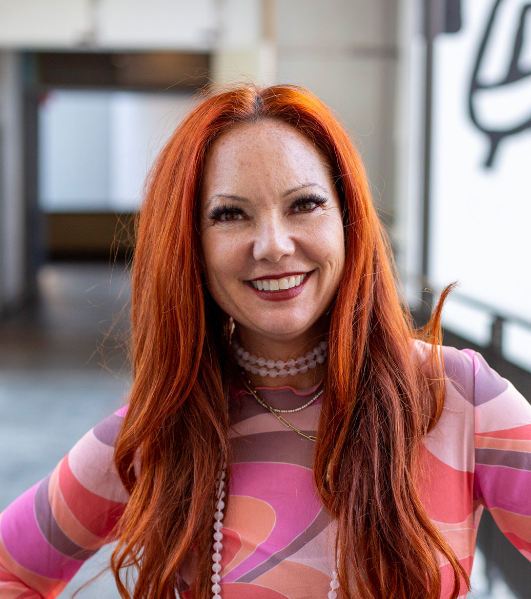 Woman smiling with long red straight hair wearing a pink and purple top with a pearl choker necklace.