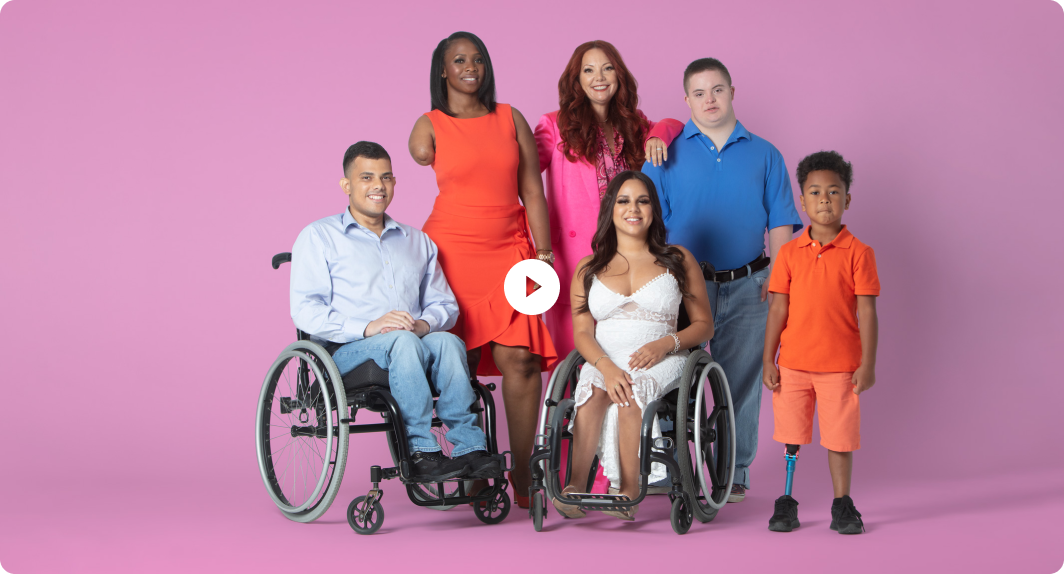 Group of models posing, with man in a wheelchair in blue pants and top, woman standing in an orange dress, Mindy Scheier in a pink suit, woman in a wheelchair wearing a white dress, teen standing in a blue shirt, and boy standing in orange top and shorts. Start video