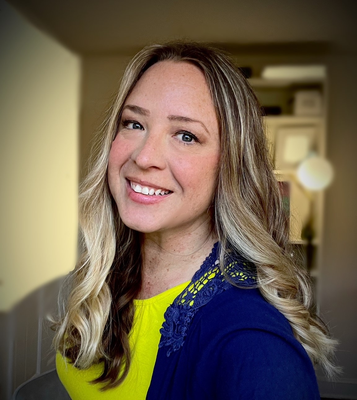 Jennifer has long blond-brown hair and is wearing a bright yellow and blue top. She is turned slightly sideways and is smiling at the camera.